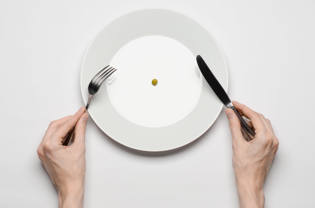 Holistic Treatment for Eating Disorders: 13 Essentials for Holistic Eating Disorder Recovery | eating disorder recovery hands holding knife and fork on a plate with green peas on a white table top eating disorder recovery - hands holding knife and fork on a plate with green peas on a white table top